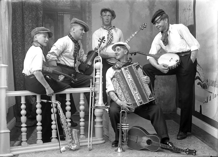 5 stycken musiker med dragspel, banjo och fiol. Framför på golvet finns saxofon, trombon, trumpet och ett fyrsträngat instrument.