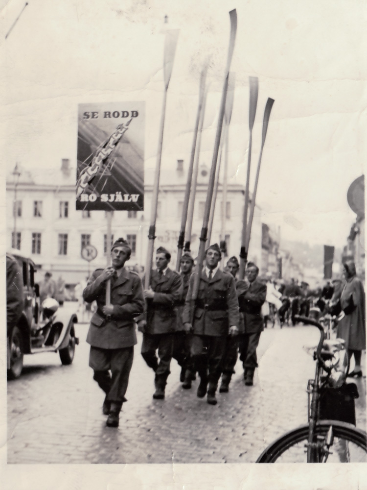 Uniformsklädda män som gör reklam för rodd gåendes på Kungsgatan i Uddevalla. En med plakat resten med åror.