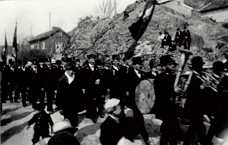 1 maj demonstration. Orkester längst fram. 4 åskådare sitter uppe i ett berg och ser på.