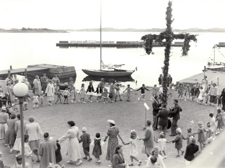 Vuxna och barn dansar kring midsommarstången. 3 mannaorkester står vid midsommarstången. Segelbåt och Gullmarn? i bakgrunden.