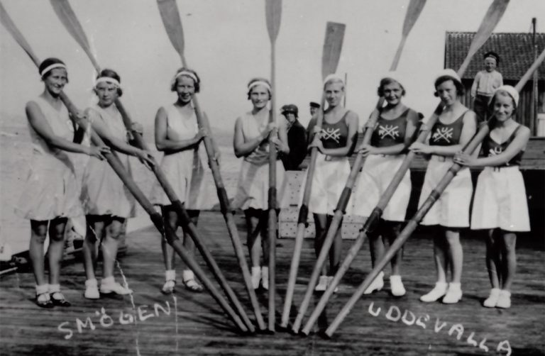 Smögen och Uddevallas kvinnliga roddlag. 8 damer som håller i var sin åra. I bakgrunden står en person och tittar på.