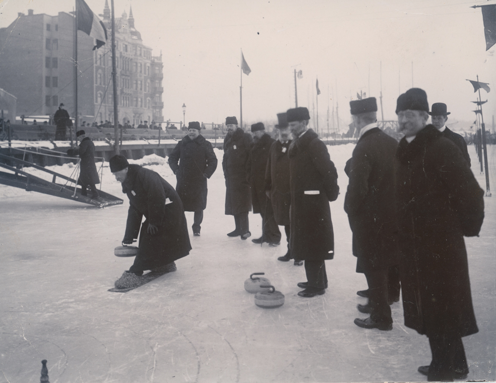 Välklädda herrar spelar curling eller ser på. En person håller precis på att släppa stenen. Flertal curlingstenar ligger på isen.