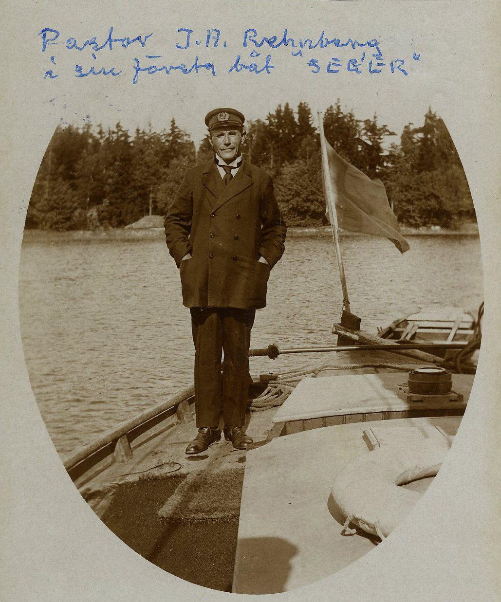 Pastor J. A. Rehnberg ombord på sin första båt Seger 1. Pastorn är klädd i skepparmössa, skepparkavaj och långbyxor. Längst bak på båten syns en flagga. På fotot är skrivet i blått blått bläck "Pastor J.A. Rehnberg i sin första båt "SEGER""