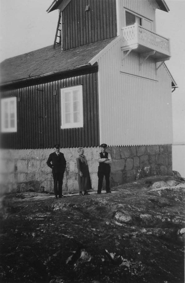 Tre personer står framför stengrunden till Varö fyr i Göteborgs södra skärgård. Fyrhuset är tvåfärgat och mot havet är huset vitt. Långt upp på den vita hussidan finns en balkong. Personerna är J.A. Rehnberg och Fyrmästare Björk och dennes hustru.