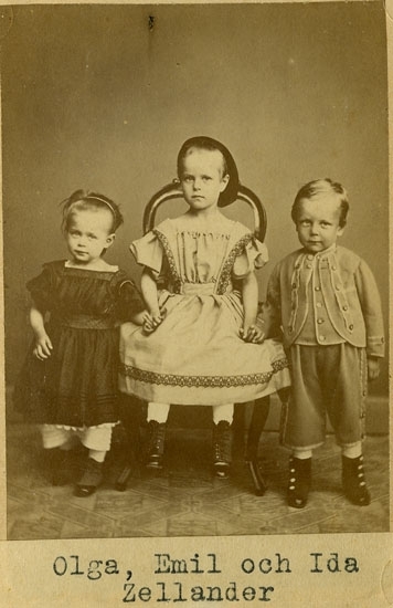 Barnen Zellander, Olga, Emil och Ida i sina bästa kläder står respektive sitter uppradade för fotografering. Barnen håller varandra i händerna. Barnen har ett utryck som kan beskrivas som allvarsamt samt spjuveraktigt.