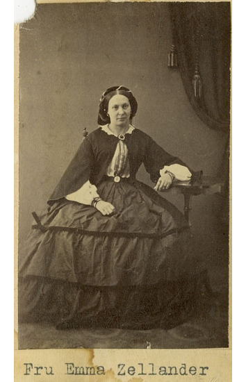 Porträtt av Fru Emma Zellander. Fru Zellander med allvarsam min sitter finklädd på en stol, vänster underarm ligger på ett bord.