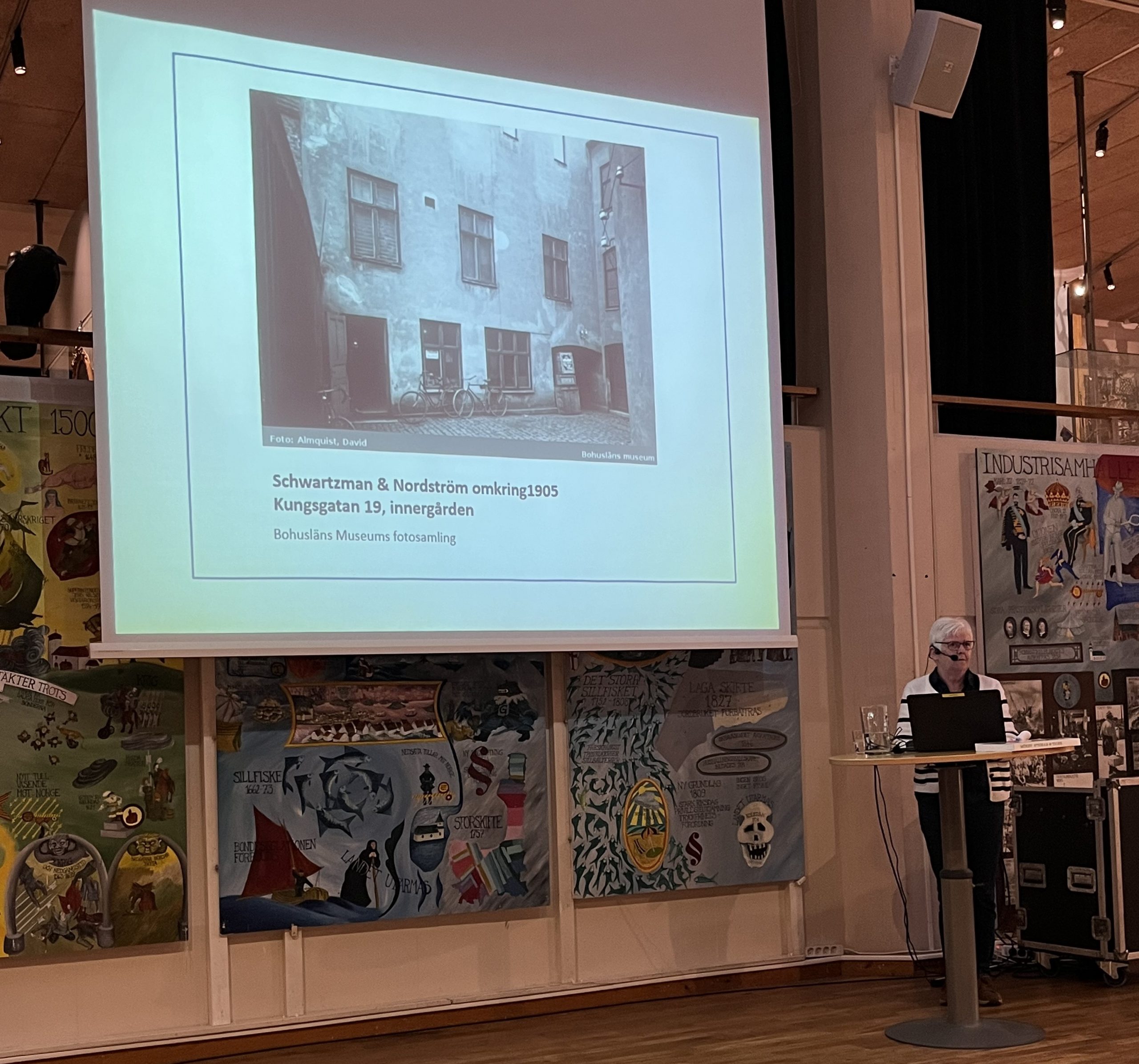 Närbild av föredragshållaren och en "slide" på duken. Sliden innehåller en bild på en byggnad och under den texten Schartzman & Nordström omkring 1905 Kungsgatan 19, innergården. Bohusläns Museum fotosamlingen.