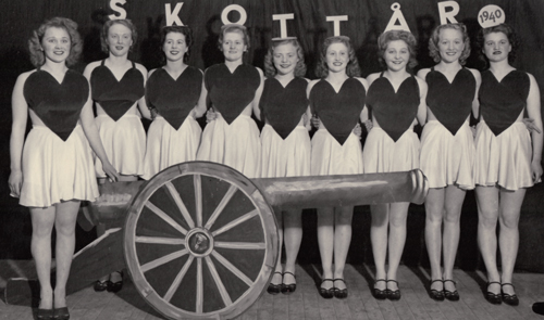9 stycken revyklädda kvinnor. Kvinnan längst till vänster står lite framför de andra och håller handen på en kanon gjord av papp. Kanonen står framför de andra kvinnorna. Bakom gruppen av kvinnor finns en ridå med texten SKOTTÅR och en rund ring med årtalet 1940 i. Bilden är svartvit.