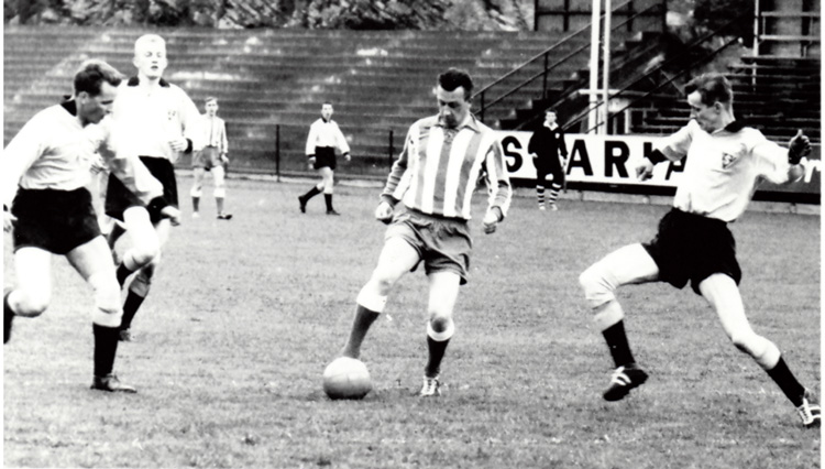 Fotbollsmatch. Spelaren mitt i bilden blir angripen av tre motståndare som försöker ta bollen ifrån honom. I bakgrunden fler spelare, en domare och läktare. Bilden är svartvit och från mitten av 1900 talet.