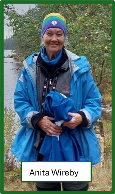 Kvinna som står vid kanten av en sjö hållandes ett par regnbyxor i händerna. På huvudet har hon en flerfärgad mössa som går i nyanserna lila till gult. Hon har flera lager lager kläder på sig och längst ut är en blå skaljacka. Benen skyms av texten Anita Wireby