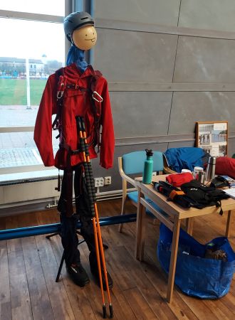 Docka utklädd till person som ska beträda en is på ett vattendrag. Dockan är klädd i hjälm, röd anorak, svarta byxor och kängor. Dockan har en ryggsäck på ryggen och isdubbar på framsidan. Mot mellangärdet på dockan lutar ett par stavar. Till höger i bild står ett bord med blanda annat en termos och vattenflaska.