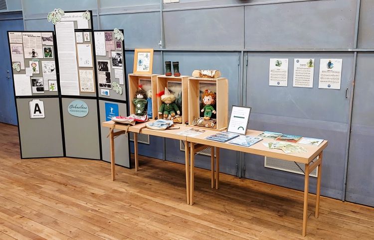 Station på en utställning. Från vänster Bohusläns föreningsarkivs skärmvägg med material om Friluftsfrämjandet. Till höger om skärmväggen står ett bord med bland annat tre lådor stående på innehållande var sin Skogsmulle. På väggen bakom bordet sitter tre papper i A4 storlek uppsatta på väggen med noter på.