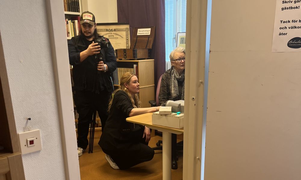 Tre personer befinner sig i ett rum som är ett arkiv. Två av dem tittar till höger i bild och den tredje som håller en kamera tittar till höger i bild.